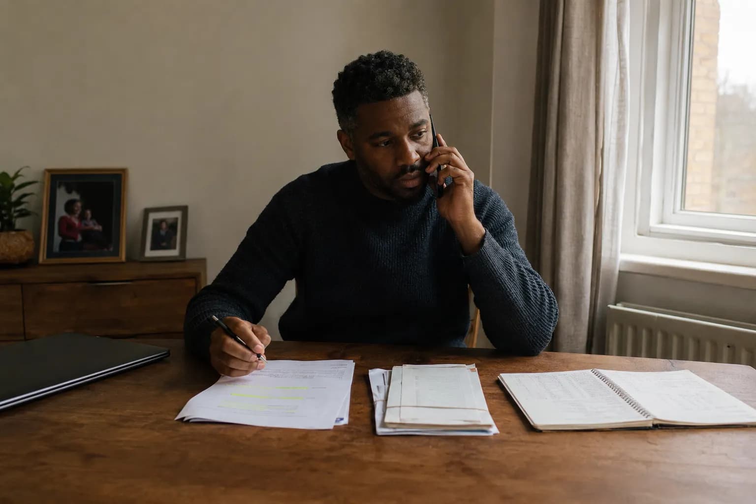 A man at his dining table on the phone to a mortgage adviser, a highlighted printout, a banded folder of older overseas statements and an open ledger laid out in front of him.