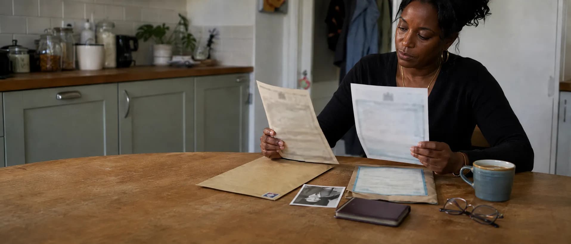 Three birth certificates of different ages spread across a kitchen table beside an old black-and-white family photograph and an opened envelope, with a woman comparing two of the certificates.