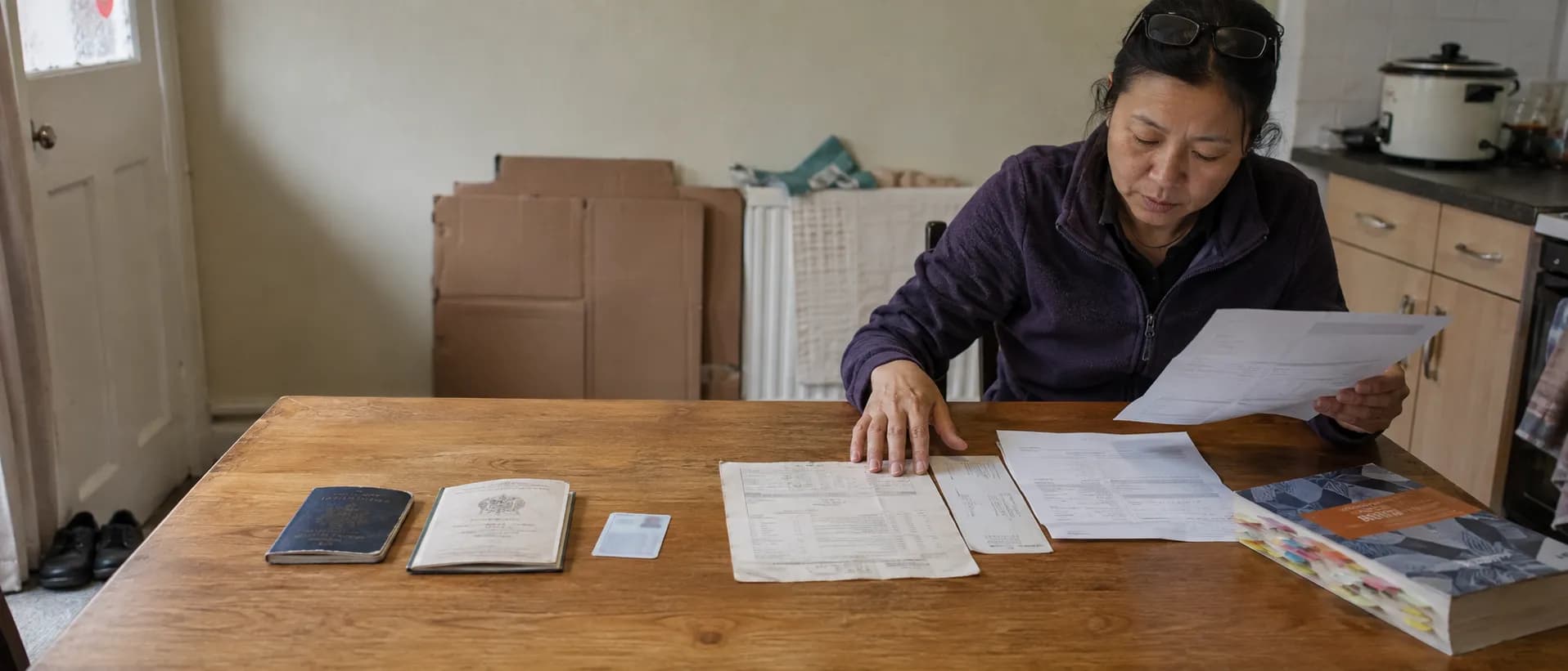 A woman at her kitchen table in a rented British house a few months after arriving, comparing two bank statements, with a BNO passport, a certificate of registration, a biometric residence permit and a study guide spread across the table.