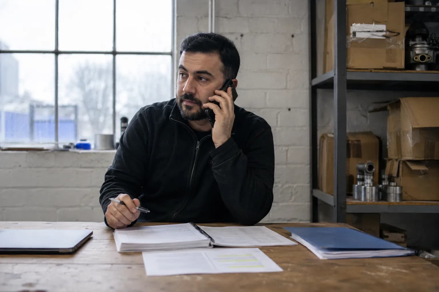 A founder on a phone call at a workbench desk in a small rented business unit, a bound set of company accounts open in front of him beside a highlighted accountant’s letter and a separate folder of personal bank statements.